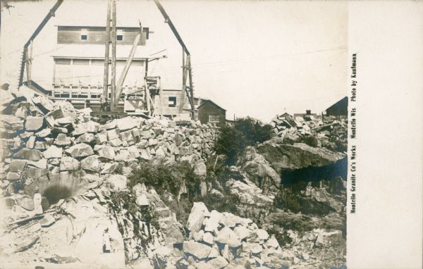 A workman is standing on the platform of a wooden derrick at the granite quarry. A two-story wood frame building is behind the derrick. Large granite blocks are supporting the upper edge of the quarry pit. Caption reads: "Montello Granite Co's Works, Montello, Wis."