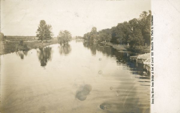 Trees are reflected in the calm waters of the Fox River just below the lock and dam at Montello. Fences are on each side of the river. The printed inscription reads: "Down Fox River at foot of Lewis' Hill. Montello. Wis. Photo by Kaufmann."