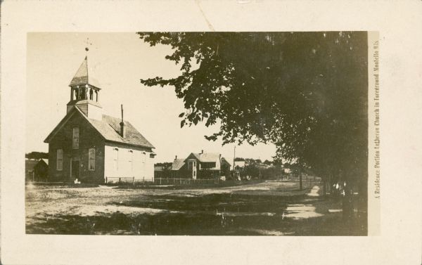 A small church with open bell tower stands along Barstow Street (now East Montello Street/Highway 23) with houses in the background. Trees along the right side of the street partially shade the unpaved sidewalk. The printed inscription reads: "A Residence Portion Lutheran Church in Foreground, Montello, Wis."