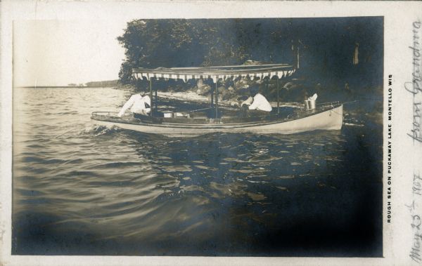 View across water towards two men sitting in a small launch with a canopy near the shore. A fishing pole is resting on the foredeck. There is a small wave in the foreground. The printed caption reads, "Rough Sea on Puckaway Lake, Montello, Wis." The written message contains the date, May 23rd, 1907, and is signed "from Grandma."