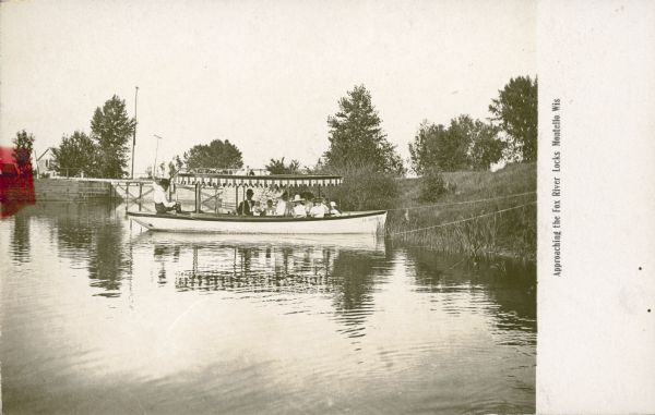View across water towards a motor launch with striped canopy moored along the shore just above the Fox River Lock on Buffalo Lake. A finely dressed party of two men, several women and children pose aboard the boat. The boat name is painted on the front right: "El Modelo." There are two men on the shore in the background; the railing of the bridge over the lock is in the far background. The caption reads: "Approaching the Fox River Locks, Montello, Wis."