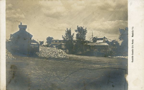 A view, looking east, of the grounds and buildings of the Montello Granite Company. On the left, a train car with "Detroit Southern" painted on it stands on tracks near a pile of uniform cut granite paving blocks. In the background, right, is the Catholic church which was later relocated for the quarry's expansion. Caption reads: "Montello Granite Co's Works, Montello, Wis."