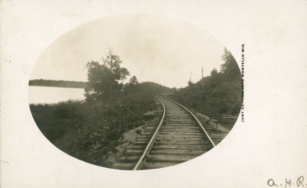 A view, framed with an oval mask, of the Wisconsin Central spur line tracks along the north shore of Buffalo Lake at Montello. The spur connected with the Wisconsin Central main line near Packwaukee. The printed caption, "Just Approaching Montello Wis." suggests the photograph was taken from the rear of a rail car as it approached the Montello depot.