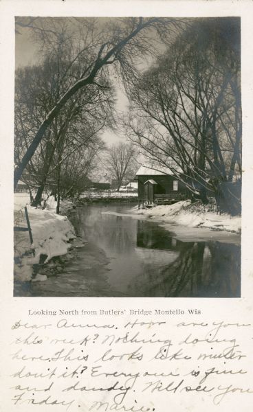 A view captioned "Looking North from Butlers' Bridge, Montello, Wis." features a shed with a snow covered roof reflected in the Montello River. There is ice near the riverbanks and snow covering the ground. In the background are sheds and the rear walls of properties that face West Montello Street. The handwritten message reads: "Dear Anna. How are you this week? Nothing doing here. This looks like winter don't it? Everyone is fine and dandy. Will see you Friday. Marie."