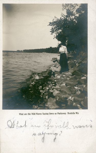 Sisters Lucy, left, and Jessie Underwood watch small waves on the shore of Lake Puckaway. Both are wearing large brimmed hats. There is a boat in the far background near the forested shoreline. The printed caption reads: "What are the Wild Waves Saying Down on Puckaway, Montello, Wis." The caption refers to a chapter title in Charles Dickens' novel "Dombey and Son" which had inspired popular songs and paintings in the Victorian era.