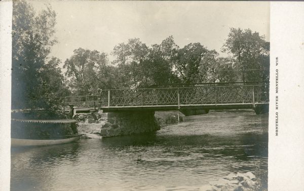 View across water towards a man holding a long pole posing on the stern of a launch near the Montello River bridge. The launch has a canopy with a scalloped edge. The bridge has a metal rail and supports and rests on an abutment made from cut blocks of granite from the local quarry. The Montello River joins the Fox River just downstream from the bridge. Caption reads: "Montello River, Montello, Wis."