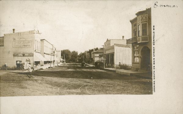 The printed inscription identifies this scene, "Looking South on Main Street, Montello, Wis." A woman is standing on the sidewalk at left near a parked buggy and wagon. There two-story brick building on the left with painted signage on the wall is O.E. Kehlet's Montello Mercantile Co. store. Other commercial buildings with large awnings line that side of the street. Toward the far end of the street, on the right, is the Marquette County Courthouse, a two-story building with a hipped, standing seam metal roof. A hanging sign identifies the Tremont House hotel, with its open porch and covered artesian well. There are two buggies in the street on the right.