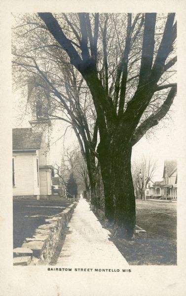 The printed inscription identifies this view of "Bairstow [sic] Street, Montello, Wis." It is a view down a sidewalk towards the Methodist Church on the left, and the steeple of St. John the Baptist Catholic Church in the far background. There is a low stone wall along the wooden sidewalk lined with mature trees on the right, with houses on the right side of the street.