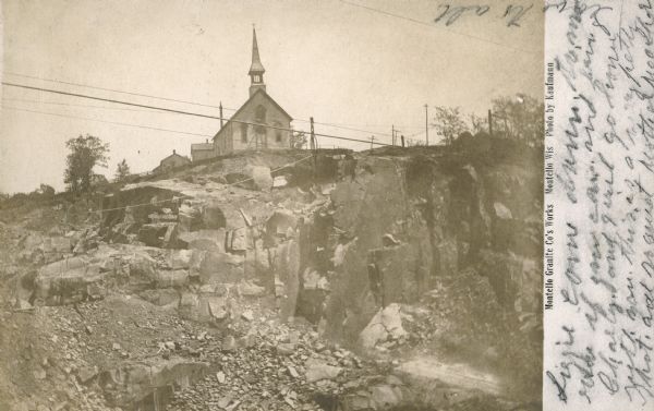View from below of a small wooden church building standing at the edge of the Montello quarry. In the foreground cables stretch over the quarry hole. The printed inscription reads, "Montello Granite Co's Works, Montello, Wis. Photo by Kaufmann." In handwriting is the message, "Lizzie, come down tomorrow if you can and bring Charly and I will go home with you. This is a very pretty spot. and so quiet. Mother [unreadable] Love to all." The church building was moved shortly after this photograph was taken to allow for expansion of the quarry. A larger Catholic church was built in a different location in 1904.