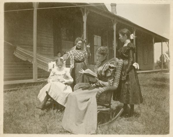 Two women and three girls posing in front of the Hotel on the Hill. Blanche Richter is seated at left and holds a cat on her lap. The woman standing next to her is unidentified. Lucy Underwood is sitting in a hammock on the porch behind the others. In the right foreground, sitting and holding a book in a wicker rocking chair, is Mrs. Charles Richter; her elder daughter Ethel May stands on the right.  