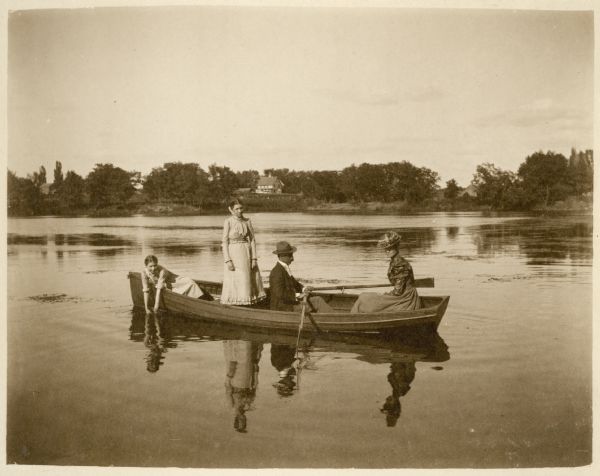 View across water towards Charles S. Richter rowing a boat on the mill pond, now Montello Lake. With him are his wife, sitting at right, his daughter Ethel May, standing, and younger daughter Blanche, sitting on far left with her hands in the water. A large barn is on the far shoreline among trees.