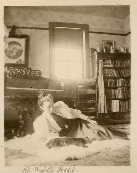 A young woman, Jessie Underwood, reclining supported on one elbow on a fur rug in front of a piano. In front of her lying on the rug is a cat. Light comes in through a window with a partially drawn shade. On the right is a bookcase crowded with books, and a stand with shelves holding periodicals next to it. A framed print (J.F. Herring's Three Pharaoh's Horses) hangs on the wall above the piano.  