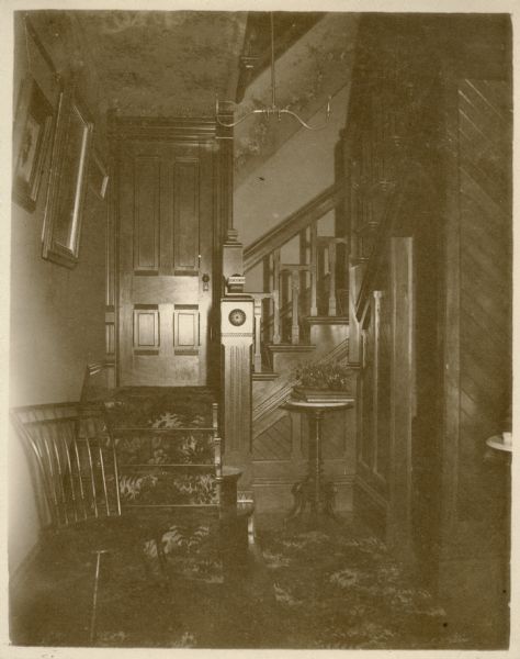 Interior view of Oakwood, the residence of E.W. Underwood showing a wood paneled U-shaped staircase. There is a chair in the foreground, and behind it is a door on the landing. Between the newel post and a doorway tucked under the stairs on the right is a plant stand holding books and two small vases with berries and foliage. A two armed gas chandelier hangs overhead. There are framed prints on the left wall, and wallpaper is on the ceiling and as a frieze on the walls.