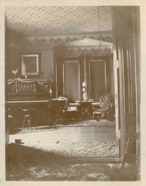 An interior available light photograph, through a doorway, of the parlor in the home of E.W. Underwood. An oil lamp is sitting on a table flanked by two chairs in a bay window. On the left is an upright piano with a fringed scarf as a covering, with two small vases on top. The one on the left holds five roses, the right holds an arrangement of cranberry stems with berries. The ceiling is wallpapered and there is a wallpaper frieze on the walls.