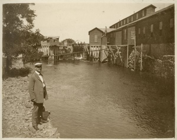 A well-dressed man is standing on the bank of the Montello River just below the dam, which was owned and operated by the Montello Granite Company. On the opposite side of the river are the Granite Company buildings, including the long polishing shed. The printed description reads, "Polishing Shops — Dr. Lange of Pittsburg [sic], Penn." J. Chris Lange was the Dean of the Medical Department of Western University of Pennsylvania (later the University of Pittsburgh) in the early twentieth century.
