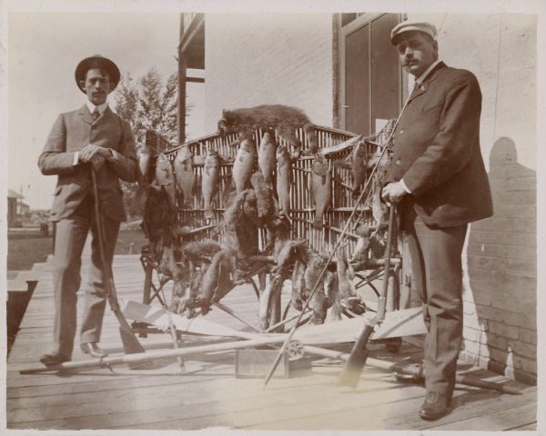 G.W. Roskie, left, of South Dakota, and Dr. Lange of Pittsburgh, Pennsylvania pose with their rifles on the porch of the Montello House Hotel. Between them is a rattan settee loaded with game including squirrels and a raccoon, and with numerous fish including gar, pike, and bass. Their fishing rods are propped against two crossed oars lying across a box on the ground. Roskie and Lange are dressed in suits.