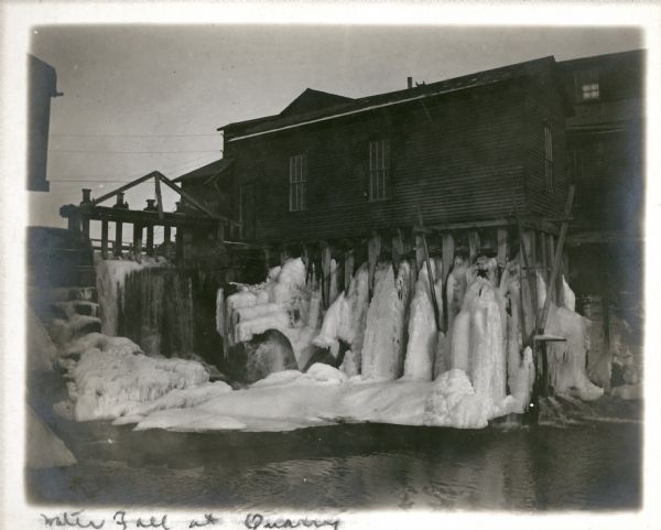A winter view of the dam, left, on the Montello River and ice formations under the shed on the right. The polishing shed of the Montello Granite Company is in the background.