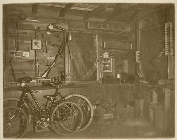 An available light photograph of the interior of the workshop of E.W. Underwood. Two bicycles lean against a workbench. There are various hand tools hanging on the walls, a fly sprayer on a shelf, and a rifle leaning against the window frame.
