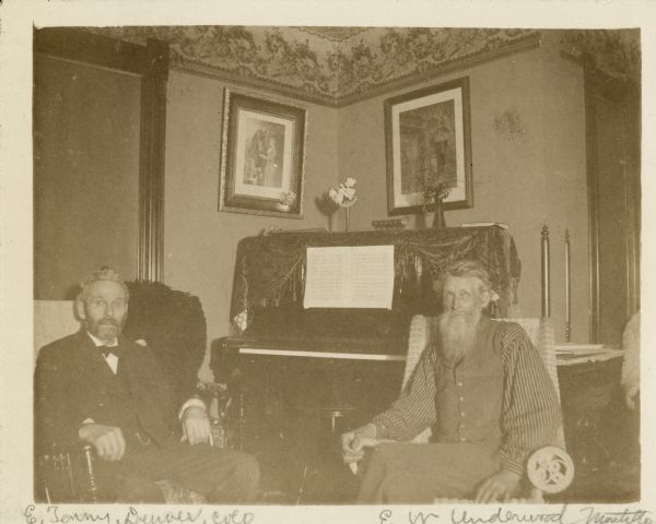 Two gentlemen, identified as E. Tenny, of Denver Colorado, left, and E.W. Underwood of Montello, Wisconsin, are posing sitting in the parlor of Underwood's home, Oakwood. Two vases sit on the piano in the background. One of the vases holds roses, the other a bunch of cranberries. Two framed prints hang from picture rail molding above the piano, and there is a wallpaper frieze above the molding.
