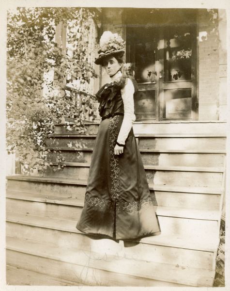Jessie Underwood, daughter of E.W. Underwood, posing on the wooden front steps of the family home. She is wearing a hat and a fancy dress with ruffled bodice and embroidered trim on the skirt. There is a leafy vine on the left and a double screened door in the background. "Jessie" has been written on the photograph.