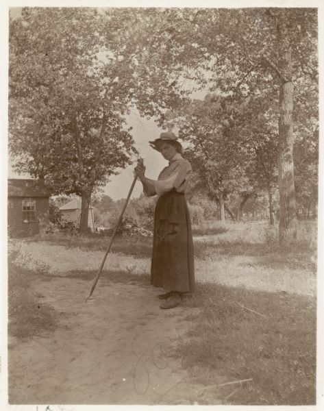 Lucy Kaufmann poses in an open, wooded area holding a hoe. She is wearing a tattered hat; her skirt and blouse are torn. There are two sheds and woodpiles in the background. Her initials, LAK, have been written on the photograph.