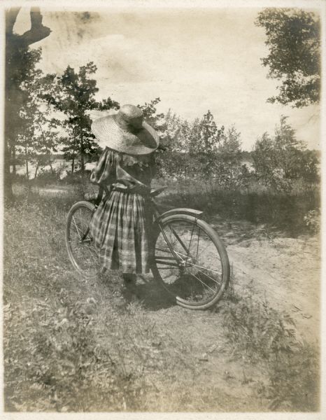 Lucy Underwood, the younger daughter of E.W. and Elenor Keyser Underwood, poses with her bicycle along a narrow sandy lane near the shore of Montello Lake. She is facing away and is wearing a large brimmed hat and a plaid dress.