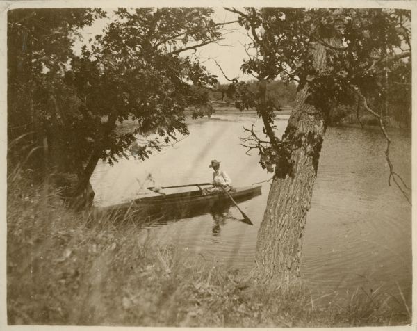 View looking down hill towards an unidentified woman who is sitting holding the oars in a skiff near the shoreline of the millpond on the Montello River, now known as Montello Lake.