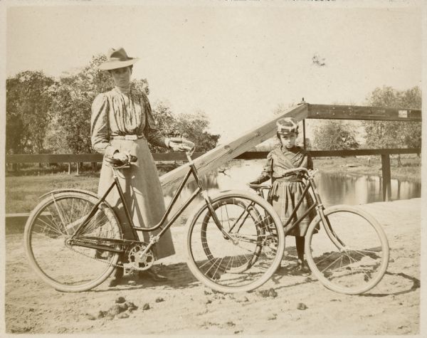 Sisters Jessie, left, and Lucy Underwood posing with their bicycles on the Montello River bridge in Montello. A man is standing in the far background on the right along the shoreline near an overturned boat.