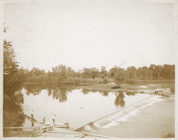 A view from the south looking over the Fox River dam at Montello. The entrance to the lock is in the background. In the foreground are two men in a boat and two men standing on the low abutment above the dam. The impoundment water forms Buffalo Lake.
