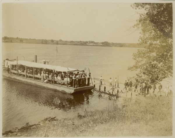 View down hill towards shoreline where a crowd fills a barge which has been fitted with benches, an awning, and American flags for an excursion on Buffalo Lake. The barge is next to a small wooden pier where several boys and men are standing. A man standing on the barge is holding a long pole. The smokestack and roof of a small rear paddle-wheel steamboat is visible behind the barge. The printed caption for the photograph reads: "Fourth of July Picnic Party."