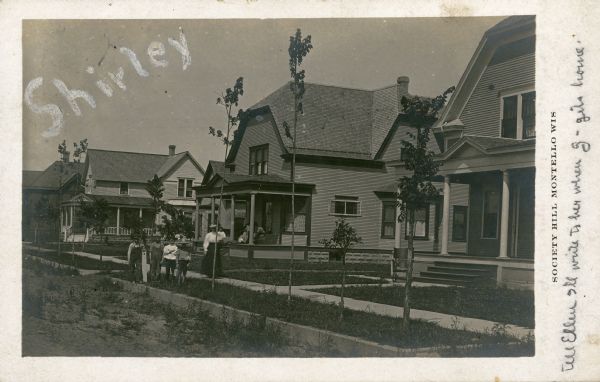 Four large, wood frame houses are seen in this view looking west along Lake Avenue. A small brick house with front bay window is visible through the open porch of the second house from the right, where three women are sitting. Another woman and four children are posing near the unpaved street. Young trees are growing between the curb and sidewalk. The printed inscription reads: "Society Hill, Montello, Wis." The sender has written on the card: "Tell Ellen I'll write to her when g - gets home." The name "Shirley" has been written across the image.