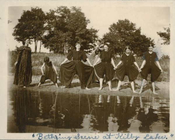 Six women and girls are reflected in the water as they model their swimming costumes on the beach. Another woman observes from behind.  The caption reads: "Bathing Scene at Kilby Lake." The women, identified on the reverse of the photograph, are, from right, "Me [Ethel M. Richter], Blanche [Richter], Katie, Miss [Lucy] Kaufmann, Lucy Underwood & Annie Caird. Mama [Mary Richter] is in the back-ground with blue glasses on." Mary Richter's husband Charles was an owner of the Montello Granite Co.  