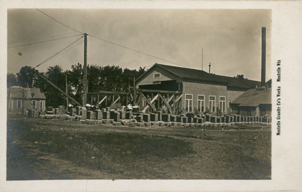 Rows of granite headstones and plinths arranged in front of buildings at the Montello quarry. A derrick is standing in the background on the left; there is a tall smokestack at right. The printed caption reads: "Montello Granite Co.'s Works, Montello, Wis."