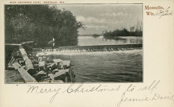 A view looking west over water toward the Fox River dam at Montello.  There is granite riprap on the left. A boat with a small sail is in the background. The impoundment waters above the dam form Buffalo Lake. A printed caption at top identifies the image as a "Miss Kaufmann Print   Montello Wis." "Merry Christmas to all. Jennie Davis" is handwritten on the front of the card, which is dated December 21. Caption reads: "Foot of Buffalo Lake."