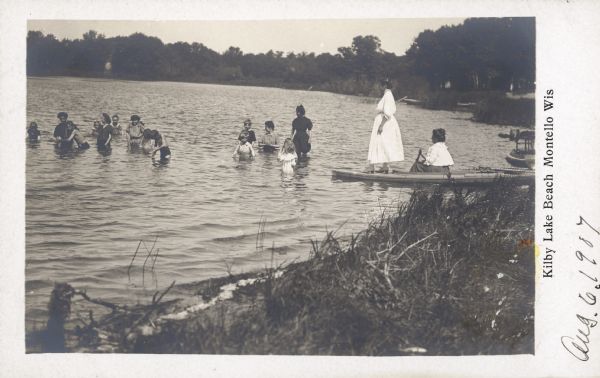 An unidentified young woman wearing a white dress is standing on the prow of a skiff watching fifteen children playing in shallow water. A second woman is sitting in the skiff holding an oar. A printed inscription identifies: "Kilby Lake Beach, Montello, Wis." "Aug. 6, 1907" is handwritten on the card.