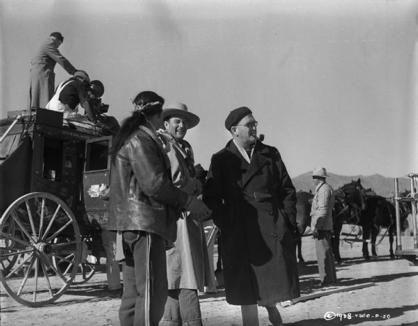 Director John Ford and John Wayne stand next to each other and an unidentified man on the set of the 1939 film Stagecoach.  A stagecoach, with a camera and two men on its roof and a team of horses, are behind the men.  Ford is smoking a pipe.