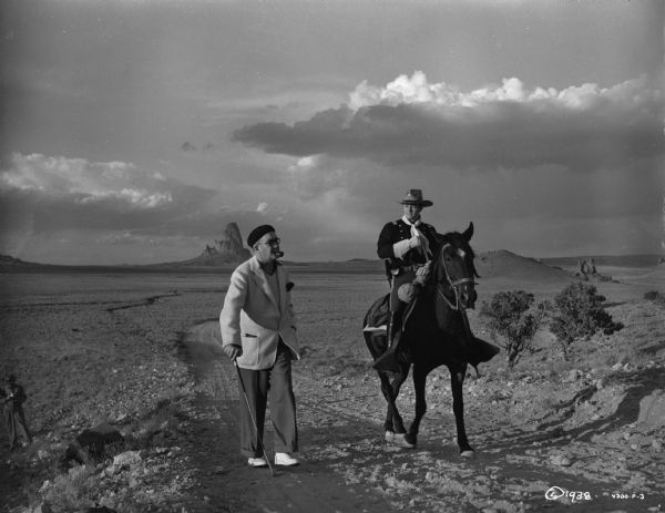 Director John Ford walks next to actor Tim Holt who is riding a horse on the set of the 1939 film Stagecoach.  Ford wears a beret and light colored jacket, is smoking a pipe, and walking with a riding crop.  There are large clouds in the sky and big rock formation can be seen in the background.