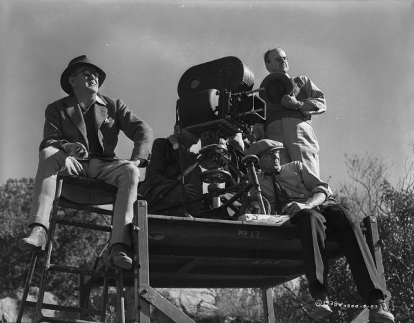 Director John Ford sits next to an elevated platform on the set of the 1939 film Stagecoach.  Director of photography Bert Glennon sits behind the camera.  Two unidentified men are seen sitting in front of and below the camera and next to it. 