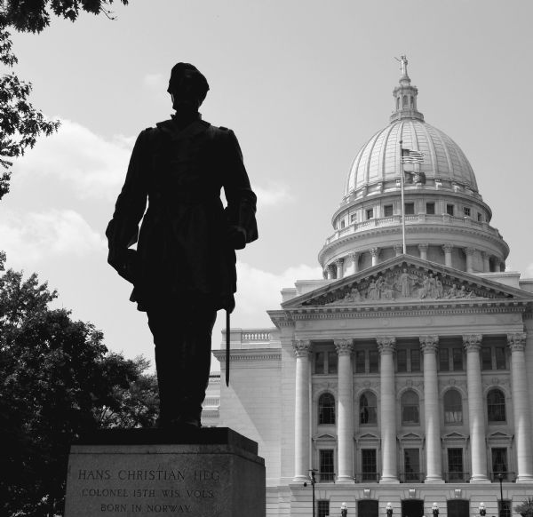View of the statue of Hans Christian Heg, with the Wisconsin State Capital in the background. Taken shortly before noon, the sculpture is silhouetted at this time of day. The base of the sculpture reads: "Hans Christian Heg, Colonel 15th Wis. Vols. Born in Norway."