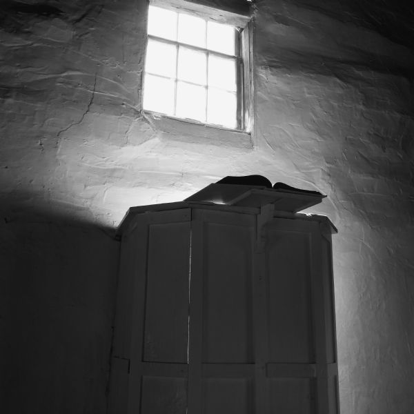 Interior view of a window sitting high up in a white lime-plastered wall, letting light into Hauge Church, a small Lutheran church built in 1852 by Norwegian immigrants. Below the window is a wooden pulpit on which is sitting an open book.