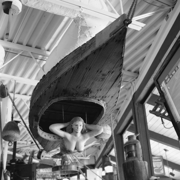 View looking up at a sculpture of a mermaid hanging from an upside down kayak suspended from the ceiling. The mermaid is wearing seashells on her breasts and has her hands placed behind her head. Buoys, ropes and pulleys, and other paraphernalia are hanging from the ceiling of the restaurant.