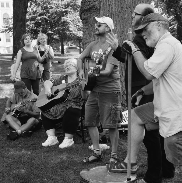 A group of men and women gathered outdoors to protest the restriction of civil rights at the Wisconsin State Capitol at noontime. Three people are playing guitars, and one man is playing a washtub bass. A woman and a man are singing. This event is known as the Solidarity Sing Along.