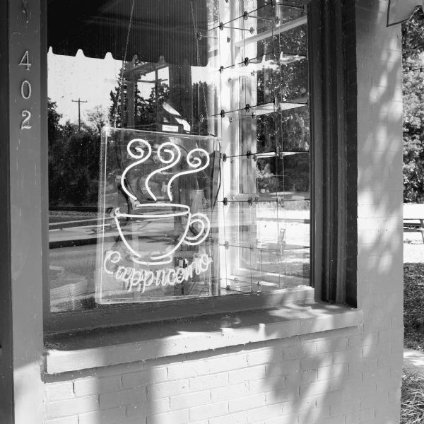 Exterior view of a neon sign hanging in the window of a coffee shop on Lakeside Street. It reads: "Cappuccino" and has a simplistic image of a coffee cup with steam rising from it. Railroad tracks are reflected in the window.