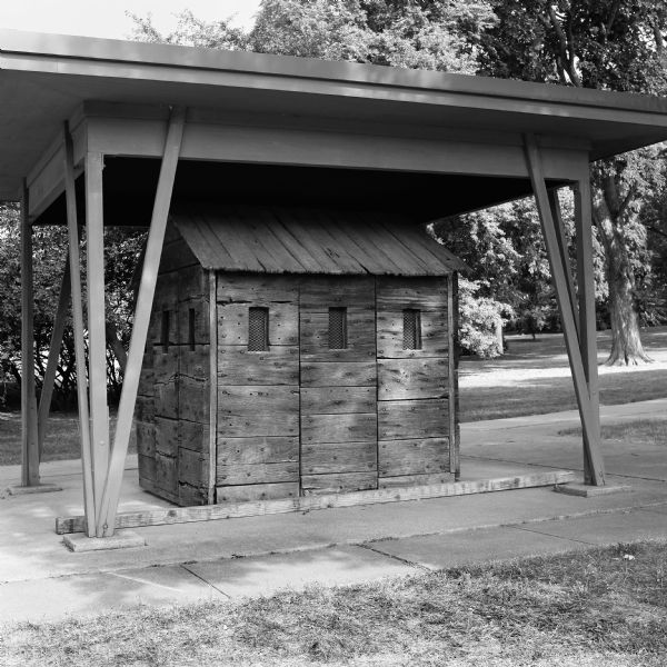 The preserved guardhouse on the site of Camp Randall under a protective awning. The guardhouse is made of wood and has metal screening in the small window openings. Trees are in the background.