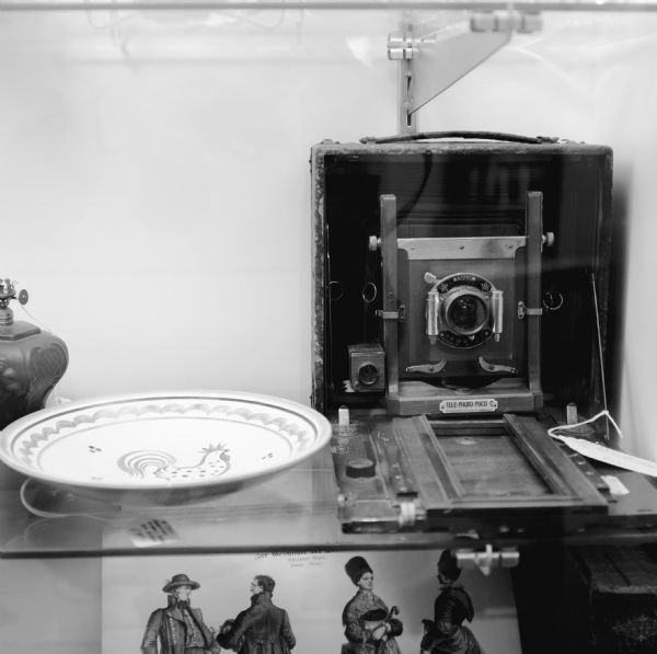 A Telephoto Poco C camera is sitting on a glass display shelf at an antique store. Next to the camera is a decorative plate, and an oil lamp. Under this shelf is a book or record with images of two men and two women. The title, in German, reads in part: "Zur Geschichte der .... Achtzehnter Bogen. Bayern. (Neuzeit)."