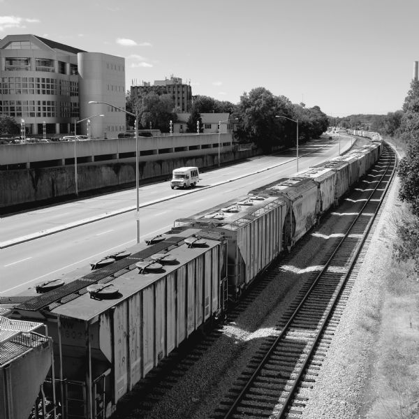 Elevated view of University Avenue from a footbridge. The University of Wisconsin Foundation and a parking lot are on the opposite side of the avenue. A Badger Bus is coming up University Avenue. On the railroad tracks in the foreground is a long train of railroad cars stretching into the distance. 