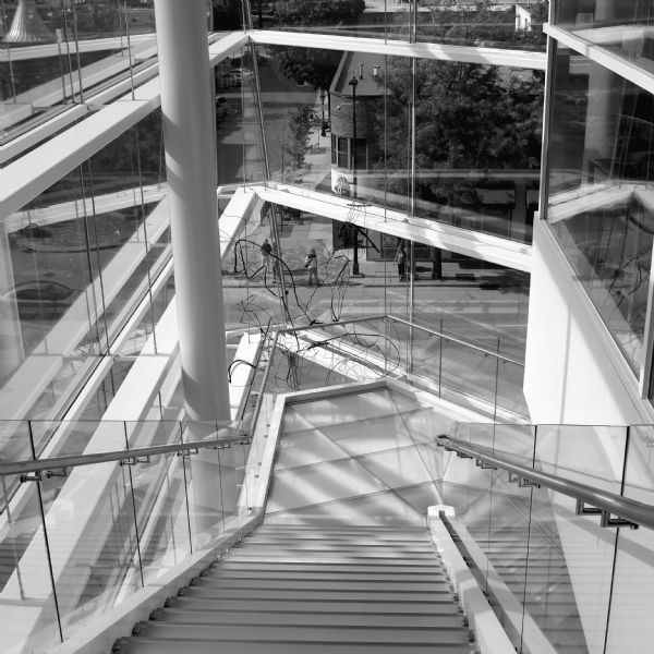View looking down at a staircase in the Madison Museum of Contemporary Art. On the ground floor is a wire sculpture that rises above the first landing of the staircase. The glass walls of the building look out towards the intersection of Johnson, State, and Henry Streets.