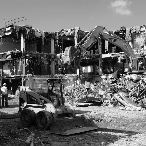 A skid steer loader is in the foreground at a demolition site, with a construction worker standing on the left surveying the site. Another construction worker is operating an excavator with a thumb attachment, dropping debris on the ground. Debris is in piles on the ground in front of the excavator. In the background are the remains of what was a three-story building, partially stripped of its exterior walls, with wires and more debris hanging from the roof and exposed floors.