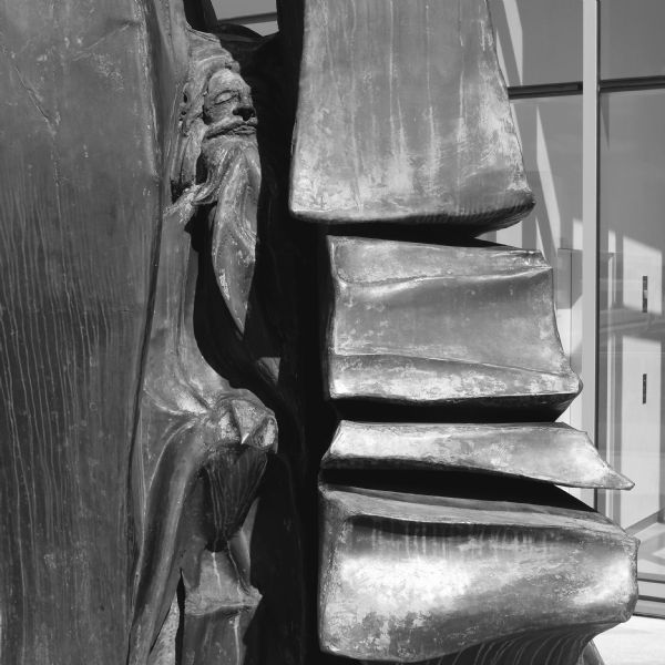 A sculpture of cast and welded metal is of a human figure with a raven, nestled in one of the crevices. This sculpture, created by O.V. Shaffer and called "Hieroglyph," is on the roof of the Madison Public Library. On the right is a large glass window or door revealing a white wall and elevator inside the building. 