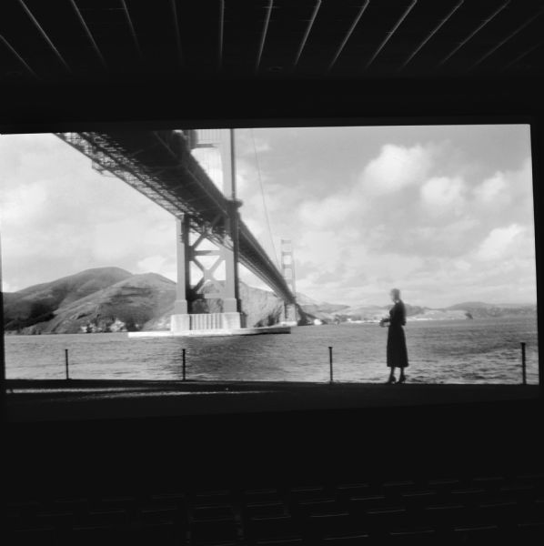 The movie "Vertigo" playing on a big screen in a darkened movie theater. The scene captured by the camera is of a woman walking along a the edge of the bay, and the Golden Gate bridge above stretching towards the opposite shoreline.